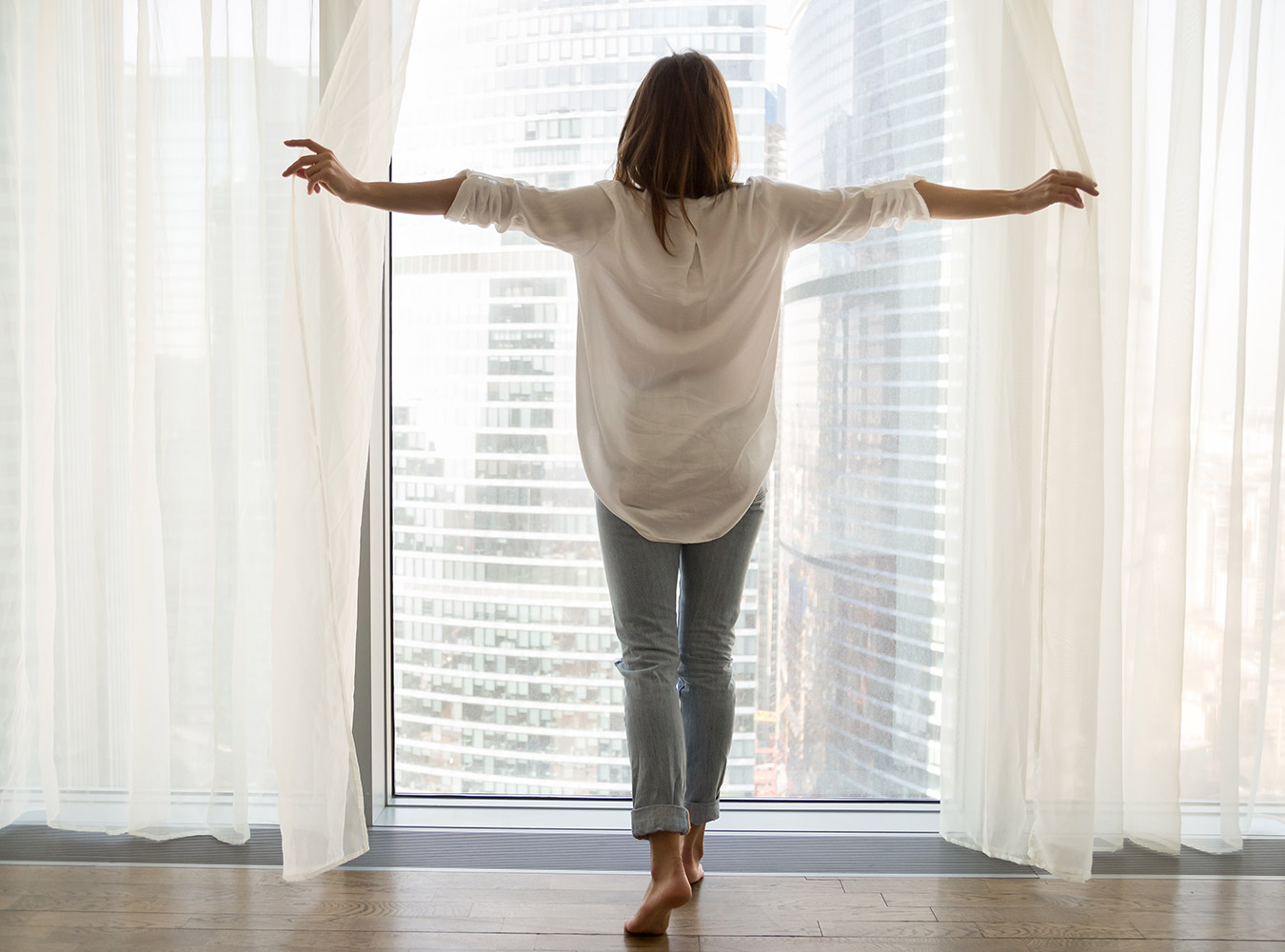 Woman stands at window with open blinds looking out over the city